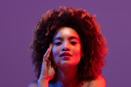 Biracial woman with curly hair, touching face in red light on purple background. Femininity, face, facial expressions, body, skin, makeup, fashion and beauty, unaltered.の写真素材