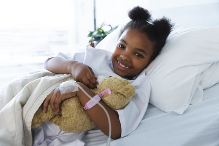 Portrait of happy african american girl patient lying on bed in patient room at hospital. Hospital, childhood, medicine and healthcare, unaltered.の写真素材