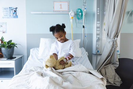 Happy african american girl patient lying on bed playing with teddy bear in patient room at hospital. Hospital, childhood, medicine and healthcare, unaltered.の写真素材