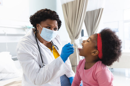 African american female doctor wearing face mask examining girl patient at hospital. Hospital, hygiene, medicine and healthcare, unaltered.の写真素材