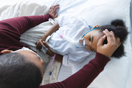 African american girl patient lying on bed with oxygen mask, with her mother at hospital. Hospital, motherhood, family, medicine and healthcare, unaltered.の写真素材