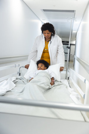 African american female doctor walking with girl patient lying on bed in corridor at hospital....