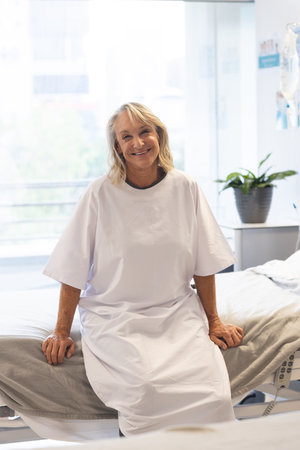 Portrait of happy senior caucasian female patient wearing hospital gown sitting on bed at hospital. Hospital, medicine and healthcare, unaltered.の写真素材