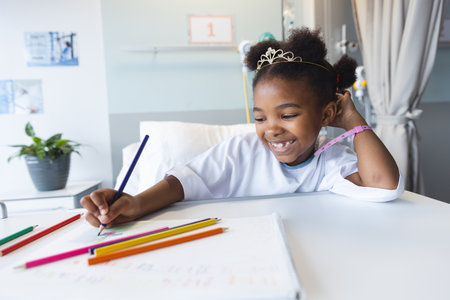 Happy african american girl patient lying on bed colouring in patient room at hospital. Hospital, childhood, medicine and healthcare, unaltered.の写真素材
