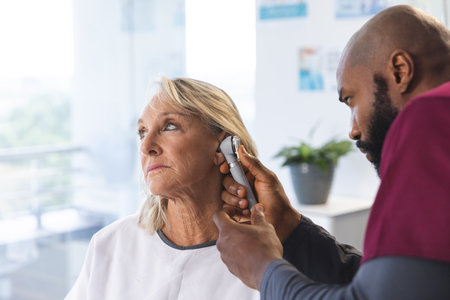 African american male doctor examining senior female caucasian patient using otoscopy. Hospital,...
