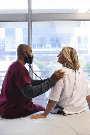 African american male doctor examining senior female caucasian patient using stethoscope at...