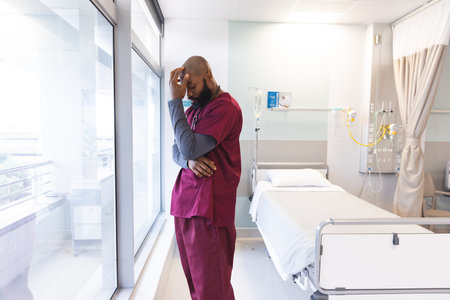 Sad african american male doctor wearing scrubs looking through window at hospital. Hospital, mental health, work, medicine and healthcare, unaltered.の写真素材