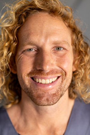 Portrait of happy caucasian male doctor with curly hair in corridor at hospital. Hospital, hygiene,...