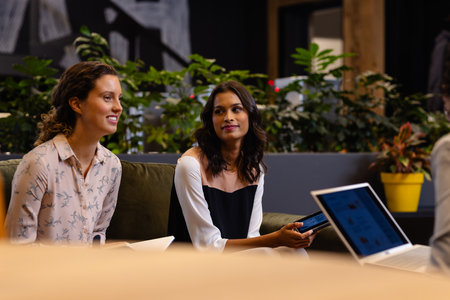 Diverse happy female colleagues in discussion using laptop in casual office meeting. Casual office, teamwork, business, lifestyle, communication and work, unaltered.の写真素材