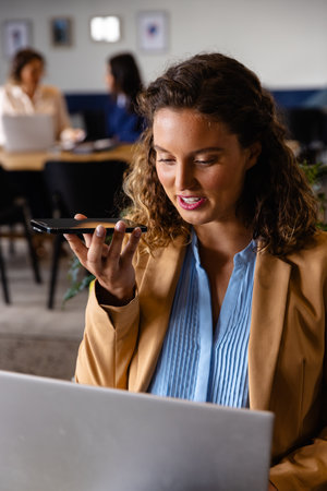 Happy caucasian casual businesswoman using laptop and talking on smartphone at desk in office. Casual office, business, lifestyle, communication and work, unaltered.の写真素材