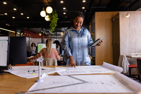 Happy plus size african american female architect using blueprints and tablet in office. Casual office, business, lifestyle, creative work, design, communication and work, unaltered.の写真素材