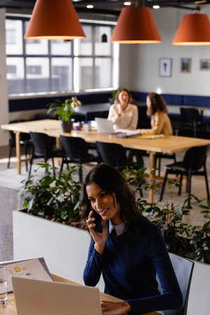 Happy middle eastern casual businesswoman using laptop and talking on smartphone at desk in office. Casual office, business, lifestyle, communication and work, unaltered.の写真素材