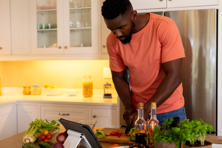 African american man chopping vegetables using tablet in kitchen. Lifestyle, food, cooking, healthy lifestyle, communication, recipe and domestic life, unaltered.の写真素材