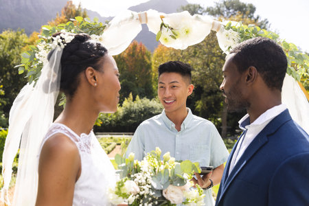 Happy diverse male officiant with bride and groom at outdoor wedding ceremony in sunny garden. Marriage, romance, summer, tradition and lifestyle, unaltered.