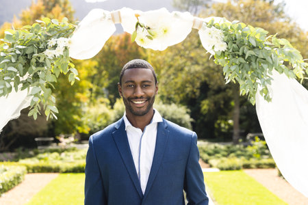Portrait of smiling african american groom standing under wedding arch in sunny garden. Marriage, romance, summer, tradition, ceremony and lifestyle, unaltered.の写真素材