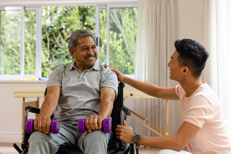 Happy diverse male physiotherapist advising and senior male patient in wheelchair using dumbbells....