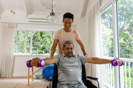 Happy diverse male physiotherapist advising and senior male patient in wheelchair using dumbbells. Physiotherapy, healthcare, wellbeing, and senior lifestyle, unaltered.の写真素材