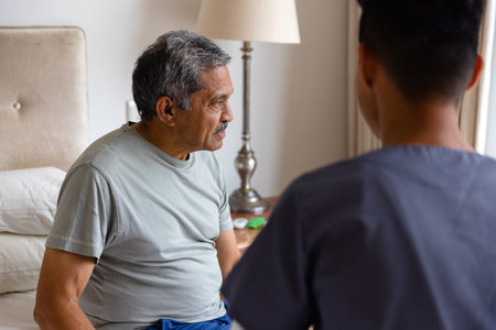Diverse male doctor and senior male patient sitting on bed and discussing at home. Medical services, check up, home visit, healthcare, wellbeing, and senior lifestyle, unaltered.の写真素材