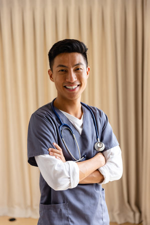 Portrait of happy asian male doctor against beige curtain. Medical services, check up, home visit, healthcare, wellbeing, and senior lifestyle, unaltered.の写真素材