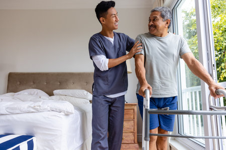 Happy diverse male doctor advising and senior male patient using crutches at home. Medical services, check up, home visit, healthcare, wellbeing and senior lifestyle, unaltered.の写真素材