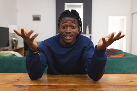 African american young man gesturing and talking over video call while sitting at table at home....