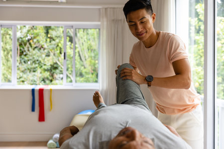 Happy diverse male physiotherapist advising and senior male patient stretching leg. Physiotherapy, healthcare, wellbeing and senior lifestyle, unaltered.の写真素材