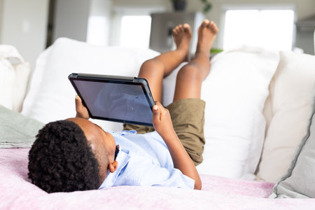 African american boy sitting on sofa and using tablet with copy space on screen. Lifestyle, childhood, communication, free time, relaxation and domestic life, unaltered.の写真素材