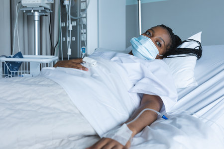 African american female patient wearing face mask, looking away, lying on bed in hospital room. Hospital, medicine and healthcare, unaltered.の写真素材