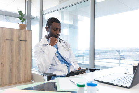 African american male doctor wearing lab coat and stethoscope, using tablet at hospital. Hospital, communication, medicine, healthcare and work, unaltered.の写真素材