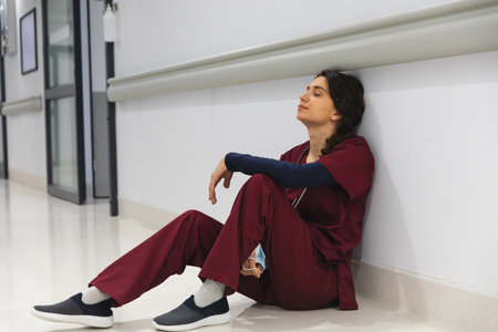 Tired caucasian female doctor wearing scrubs, sitting on floor in corridor at hospital. Hospital, mental health, medicine, healthcare and work, unaltered.の写真素材