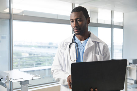 African american male doctor wearing lab coat and stethoscope, using laptop at hospital. Hospital, communication, medicine, healthcare and work, unaltered.の写真素材