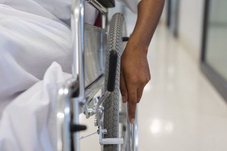 Close up of african american female patient wearing hospital gown, sitting in wheelchair in corridor. Hospital, disability, medicine and healthcare, unaltered.の写真素材