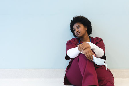 Tired african american female doctor sitting on floor in corridor at hospital, copy space. Hospital, mental health, medicine, healthcare and work, unaltered.の写真素材