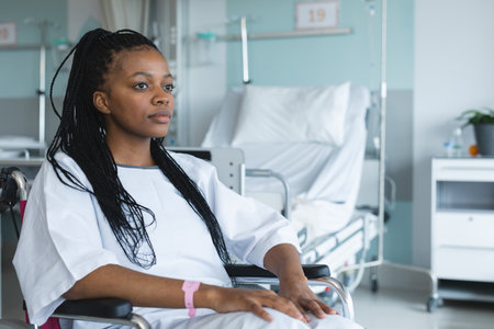 African american female patient wearing hospital gown, sitting in wheelchair in hospital room....