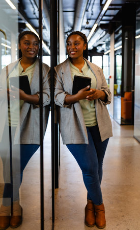 Happy plus size african american casual businesswoman holding tablet in office corridor. Casual office, communication, business, professional and work, unaltered.の写真素材