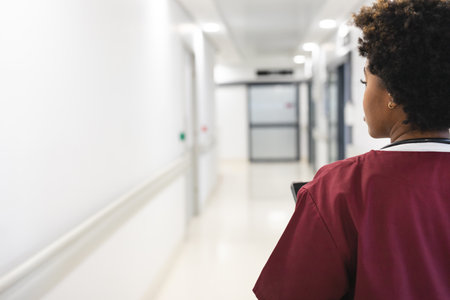 African american female doctor wearing scrubs, walking through corridor at hospital, copy space. Hospital, medicine, healthcare and work, unaltered.の写真素材