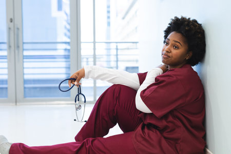 Tired african american female doctor wearing scrubs, sitting on floor in corridor at hospital. Hospital, mental health, medicine, healthcare and work, unaltered.の写真素材