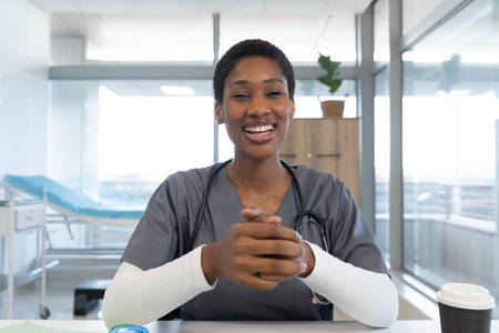 African american female doctor wearing scrubs and stethoscope, having video call at hospital. Hospital, communication, video call, medicine, healthcare and work, unaltered.の写真素材