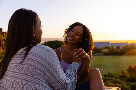 Biracial lesbian couple sitting and embracing in garden at sunset. Lifestyle, relationship, togetherness, nature and domestic life, unaltered.の写真素材
