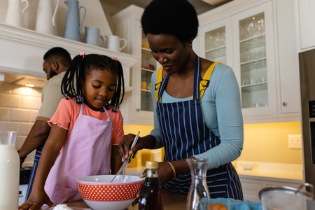 African american mother and daughter mixing batter in bowl on table in kitchen. Unaltered, lifestyle, family, love, togetherness, childhood, food, preparation and learning concept.の写真素材