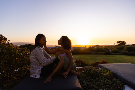 Biracial lesbian couple sitting in garden at sunset. Lifestyle, relationship, togetherness, nature and domestic life, unaltered.の写真素材