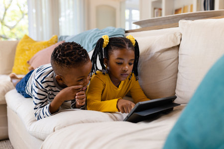African american siblings using digital tablet while lying and relaxing on sofa in living room. Unaltered, childhood, family, togetherness, relaxation, wireless technology and home concept.の写真素材