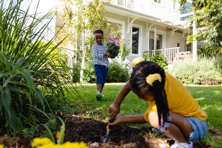 African american boy holding flower pot walking towards sister digging dirt on grassy field in yard. Unaltered, lifestyle, gardening, family, love, togetherness, nature and childhood.の写真素材