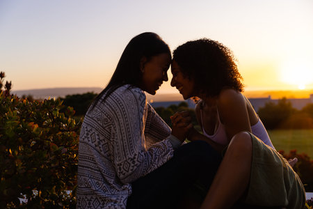 Biracial lesbian couple sitting and holding hands in garden at sunset, copy space. Lifestyle, relationship, togetherness, nature and domestic life, unaltered.の写真素材