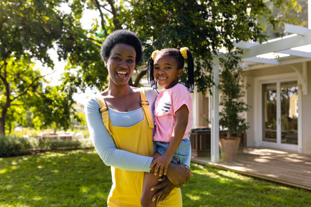 Portrait of african american cheerful mother carrying daughter while standing in yard outside house. Unaltered, lifestyle, family, love, togetherness, happy and childhood concept.の写真素材