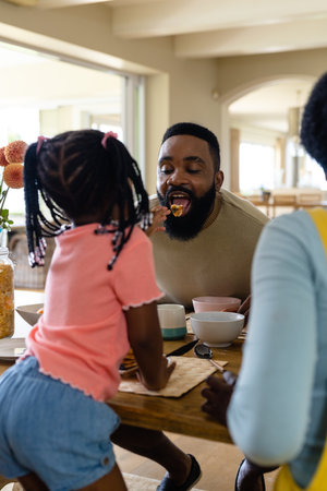 African american daughter feeding pancake to father while having breakfast at dining table. Unaltered, lifestyle, family, love, togetherness, childhood, morning and home concept.の写真素材