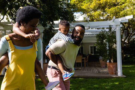 African american parents piggybacking son and daughter while walking in backyard on sunny day. Unaltered, lifestyle, family, love, togetherness, childhood, playing and enjoyment concept.の写真素材