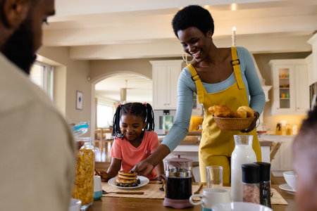 Smiling african american mother serving pancakes to daughter sitting at dining table. Unaltered,...