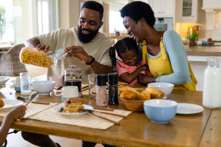 African american parents and daughter laughing cheerfully and having breakfast at dining table. Unaltered, lifestyle, family, love, togetherness, childhood, morning, food, home concept.の写真素材