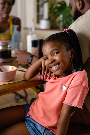 Portrait of african american girl smiling at camera while having breakfast with parents at table. Unaltered, lifestyle, family, love, togetherness, childhood, food, morning and home concept.の写真素材